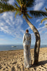 Model wearing a silver sequin blazer with a wrap style front and exaggerated shawl lapel, paired with high rise silver sequin trousers with a wide leg cut, creating a flattering, elongated, and statement making silhouette, standing on a beach with palm trees and ocean in the background.