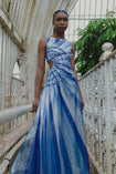 Model wearing a blue and white sunray stripe floor-length sequin dress standing on a bridge with greenery in the background