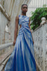 Model wearing a blue and white sunray stripe floor-length sequin dress standing on a bridge with greenery in the background