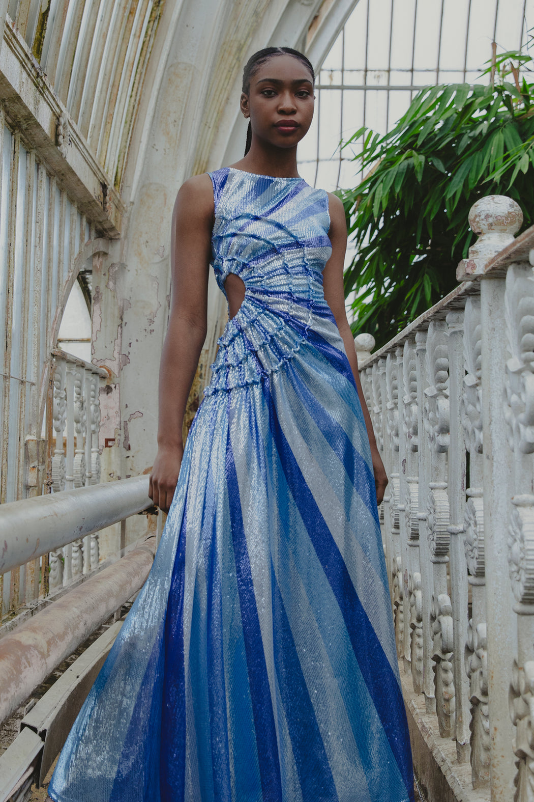 Model wearing a blue and white sunray stripe floor-length sequin dress standing on a bridge with greenery in the background
