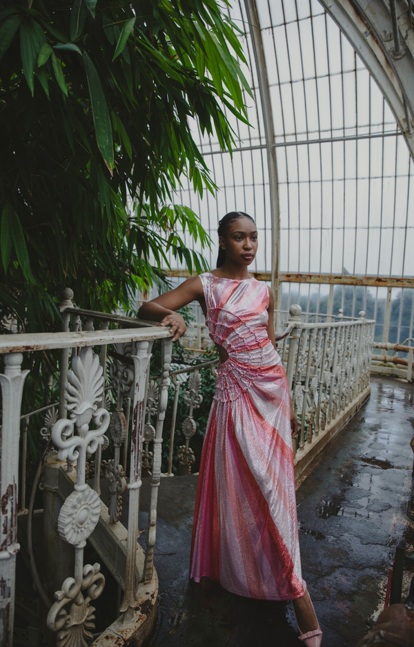 Model wearing a pink statement dress with sunray stripes, all-over shimmering sequins, intricate gathered tuck detailing, sleeveless design, round neckline, ruched bodice, and an adjustable side cut-out at the waist for a flattering, eye-catching silhouette, standing on a balcony with greenery and architectural elements in the background.