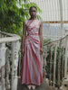 Model wearing a pink statement dress with sunray stripes, all-over shimmering sequins, intricate gathered tuck detailing, sleeveless design, round neckline, ruched bodice, and an adjustable side cut-out at the waist for a flattering, eye-catching silhouette, standing in a greenhouse.