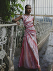 Model wearing a pink statement dress with sunray stripes, all-over shimmering sequins, intricate gathered tuck detailing, sleeveless design, round neckline, ruched bodice, and an adjustable side cut-out at the waist for a flattering, eye-catching silhouette, standing on a balcony with decorative railings.
