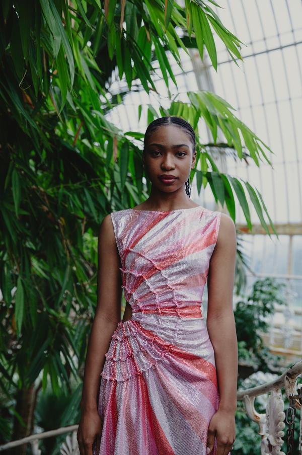 Model wearing a pink statement dress with sunray stripes, all-over shimmering sequins, intricate gathered tuck detailing, sleeveless design, round neckline, ruched bodice, and an adjustable side cut-out at the waist for a flattering, eye-catching silhouette, standing in a lush green outdoor setting.