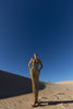 Model in a gold pleated ankle-length sequin dress with unique tuck detailing down the front with a separate bamboo jersey lace standing on a sand dune under a clear blue sky.