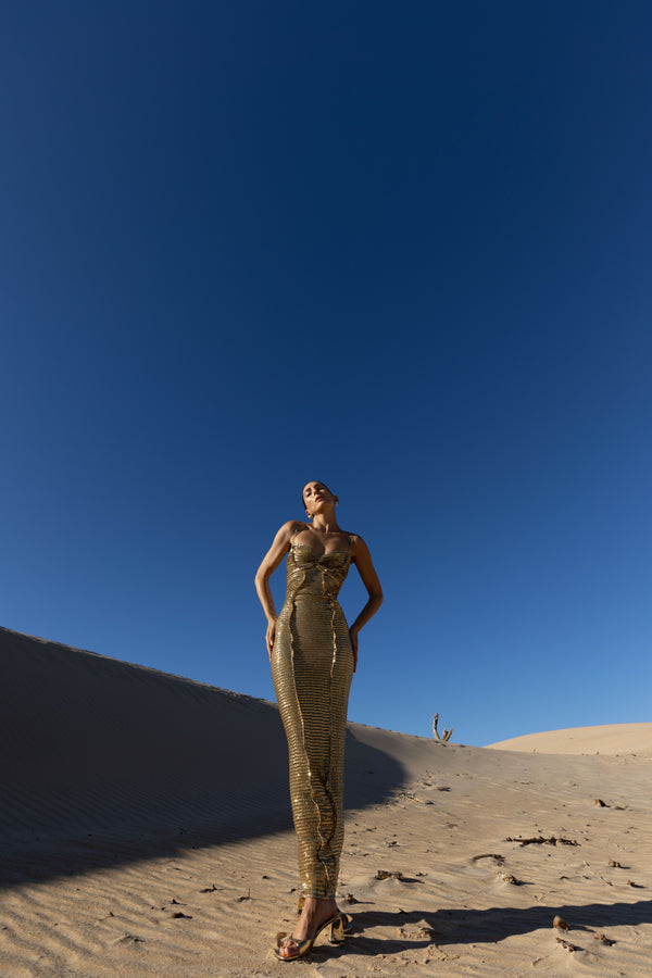 Model in a gold pleated ankle-length sequin dress with unique tuck detailing down the front with a separate bamboo jersey lace standing on a sand dune under a clear blue sky.