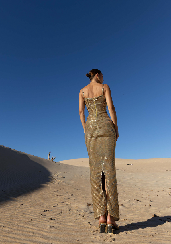 Model wearing a form-fitting gold sequin pleated ankle-length dress with thin straps and a back slit, standing on sand dunes under a clear blue sky.