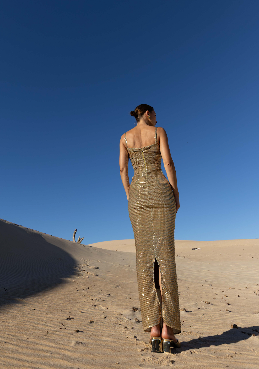 Model wearing a gold sequin ankle-length dress with thin straps and a back slit standing on sand dunes under a clear blue sky.
