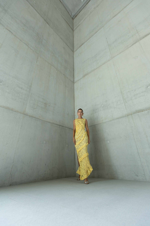 Model wearing a striking contemporary lemon-coloured floor length dress standing in a concrete room, pleated all over with statement tucks in a circular motion on an intricate French Baroque style lace, flattering the body.