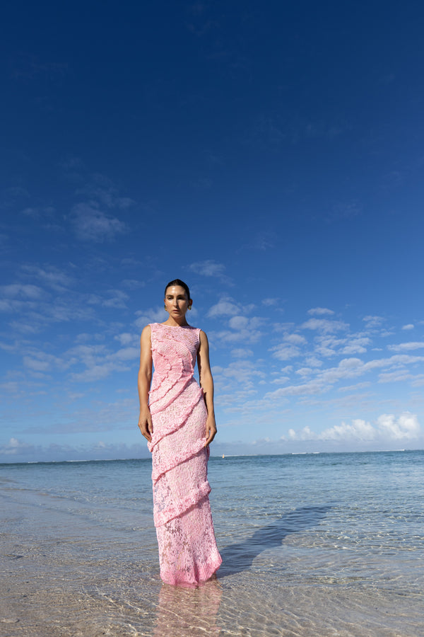 Model wearing a pink striking contemporary floor length dress, pleated all over with statement tucks in a circular motion on an intricate French Baroque style lace, flattering the body. Features a gathered panel on the side to nip in and draw focus to the waist. A slit at the side provides ease when worn, standing on a beach with clear blue sky and ocean.