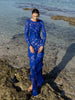 Model wearing a blue striking contemporary floor length dress, pleated all over with statement tucks in a circular motion on an intricate French Baroque style lace, flattering the body. Features a gathered panel on the side to nip in and draw focus to the waist. A slit at the side provides ease when worn. This dress has full length sleeves, standing on a rocky beach.
