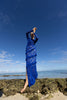 Model wearing a blue striking contemporary floor length dress, pleated all over with statement tucks in a circular motion on an intricate French Baroque style lace, flattering the body. Features a gathered panel on the side to nip in and draw focus to the waist. A slit at the side provides ease when worn. This dress has full length sleeves, standing on a rocky beach with clear blue sky and ocean.