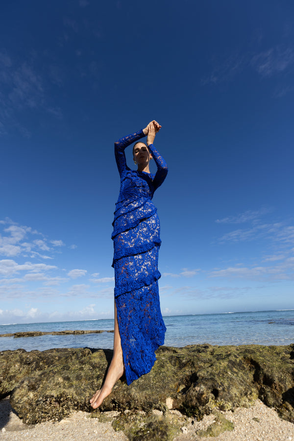 Model wearing a blue striking contemporary floor length dress, pleated all over with statement tucks in a circular motion on an intricate French Baroque style lace, flattering the body. Features a gathered panel on the side to nip in and draw focus to the waist. A slit at the side provides ease when worn. This dress has full length sleeves, standing on a rocky beach with clear blue sky and ocean.