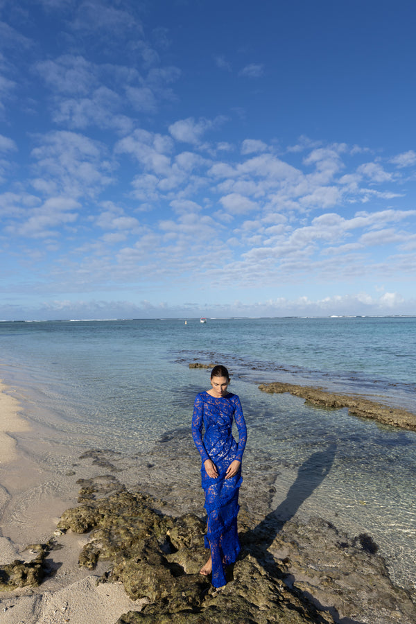 Model wearing a blue striking contemporary floor length dress, pleated all over with statement tucks in a circular motion on an intricate French Baroque style lace, flattering the body. Features a gathered panel on the side to nip in and draw focus to the waist. A slit at the side provides ease when worn. This dress has full length sleeves, standing on a rocky beach with clear blue sky and ocean.