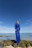Model wearing a blue striking contemporary floor length dress, pleated all over with statement tucks in a circular motion on an intricate French Baroque style lace, flattering the body. Features a gathered panel on the side to nip in and draw focus to the waist. A slit at the side provides ease when worn. This dress has full length sleeves, standing on a rocky beach with clear blue sky and ocean.