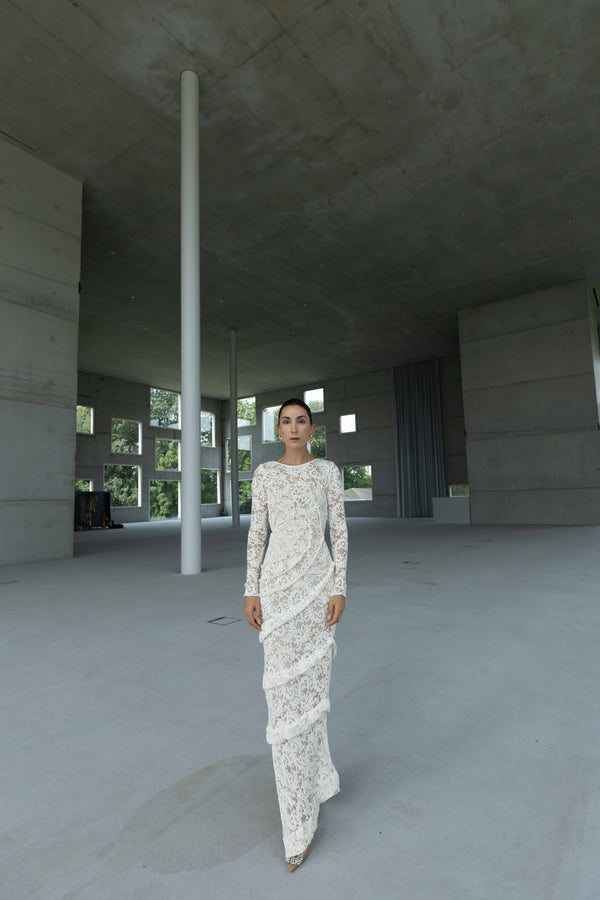 Model wearing a striking contemporary ivory-coloured floor length dress against the concrete wall, pleated all over with statement tucks in a circular motion on an intricate French Baroque style lace, flattering the body. This dress has full length sleeves, standing in a modern architectural space.