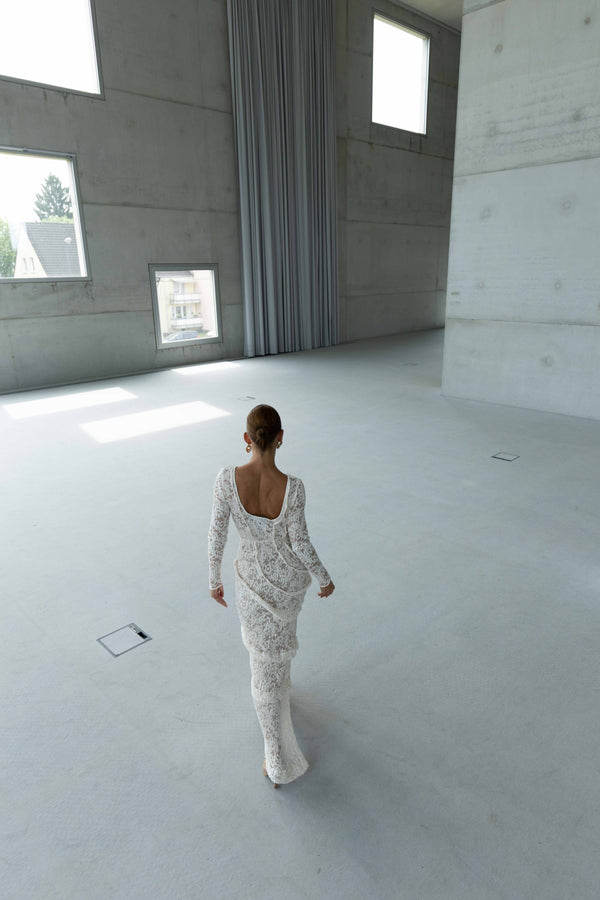A striking contemporary ivory-coloured floor length dress against the concrete wall, pleated all over with statement tucks in a circular motion on an intricate French Baroque style lace, flattering the body. This dress has full length sleeves, walking in a minimalist room with concrete walls and floor.