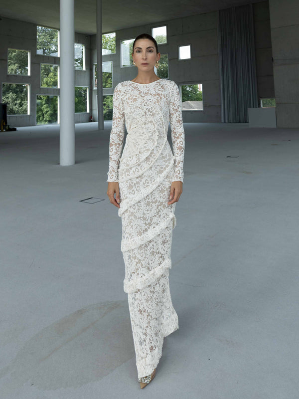 Model wearing a striking contemporary ivory-coloured floor length dress against the concrete wall, pleated all over with statement tucks in a circular motion on an intricate French Baroque style lace, flattering the body. This dress has full length sleeves, standing in a modern indoor setting with large windows.
