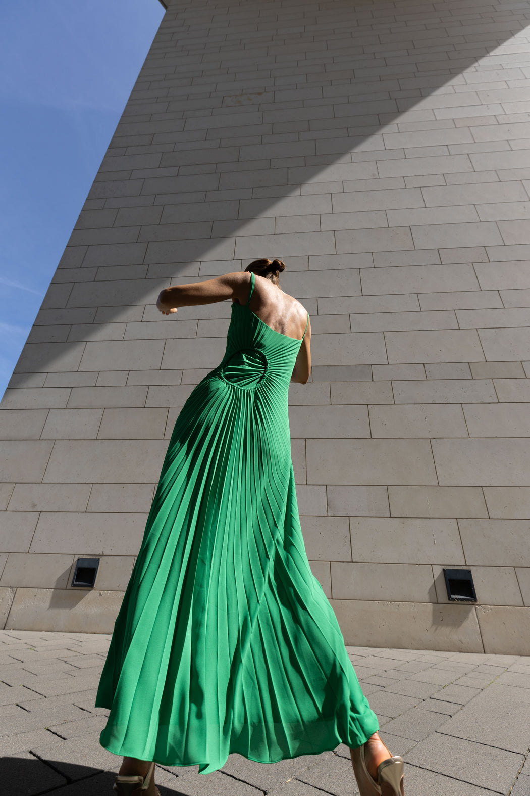 Model in a green dress pleated in contrasting directions standing against a stone wall with blue sky.