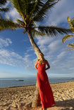 Model wearing red crafted from certified recycled georgette fabric, this piece embodies sustainable elegance with a contemporary modern design. Intricately pleated in contrasting directions, the garment creates a flattering silhouette, standing next to a palm tree on a beach with ocean and sky in the background.