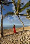 Model wearing red crafted from certified recycled georgette fabric, this piece embodies sustainable elegance with a contemporary modern design. Intricately pleated in contrasting directions, the garment creates a flattering silhouette, standing under palm trees on a beach with ocean view.