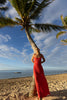 Model wearing red crafted from certified recycled georgette fabric, this piece embodies sustainable elegance with a contemporary modern design. Intricately pleated in contrasting directions, the garment creates a flattering silhouette, standing next to a palm tree on a beach with ocean and sky in the background.