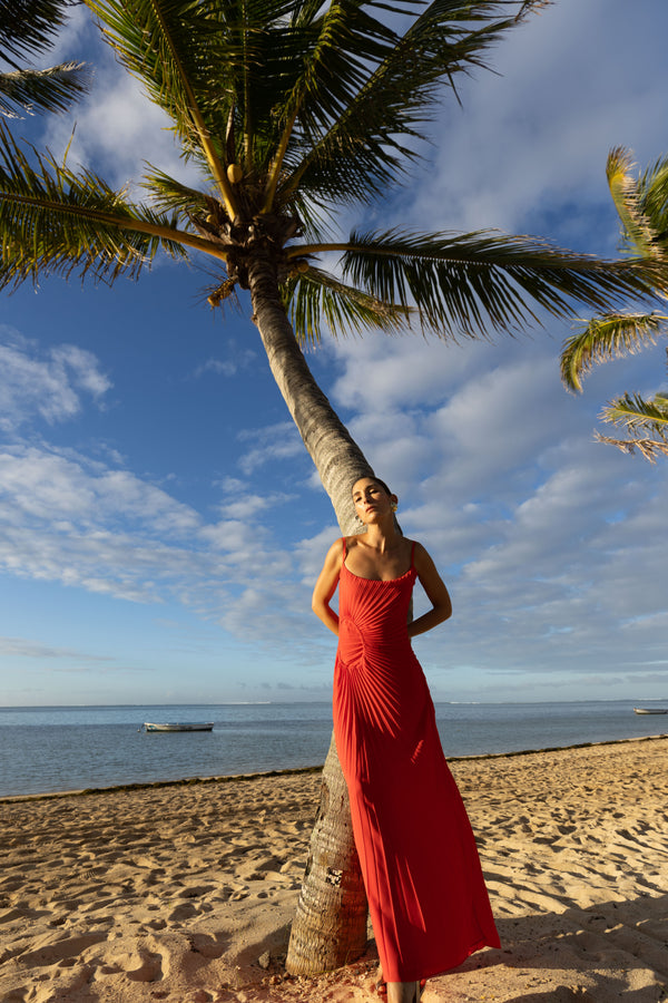 Model wearing red crafted from certified recycled georgette fabric, this piece embodies sustainable elegance with a contemporary modern design. Intricately pleated in contrasting directions, the garment creates a flattering silhouette, standing next to a palm tree on a beach with ocean and sky in the background.