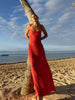 Model wearing red crafted from certified recycled georgette fabric, this piece embodies sustainable elegance with a contemporary modern design. Intricately pleated in contrasting directions, the garment creates a flattering silhouette, standing next to a palm tree on a beach with ocean and sky in the background.