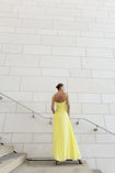 Model in a green georgette dress pleated in contrasting directions standing on a staircase against a light gray wall.