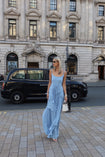 Model in a blue floor length dress pleated in a flowing waterfall design standing on a city street with a black car in the background.