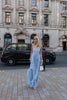 Model in a blue floor length dress pleated in a flowing waterfall design standing on a city street with a black car in the background.