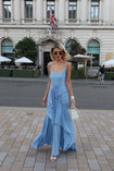 Model wearing a blue floor-length pleated dress with a flowing waterfall design, walking on a street beside a building with a UK flag
