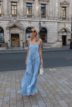 Model wearing a blue floor-length pleated dress with a flowing waterfall design standing on a city street