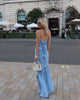 Model wearing a blue floor-length pleated dress with a flowing waterfall design. This is the back of the dress, standing on a city street.
