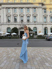 A side view photo where a model is holding a white handbag, wearing a blue floor-length pleated dress with a flowing waterfall design, standing on a city street with classical architecture in the background.