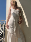 Model in a white dress wearing a floor-length dress with delicate waterfall pleating, an open neckline with adjustable straps, and a slip-on waist tie creating a flattering silhouette, standing against a wall with a blue sky background.
