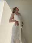 Model in a white dress wearing a floor length dress with delicate waterfall pleating, an open neckline with adjustable straps, and a slip on waist tie creating a flattering silhouette, standing against a light colored wall.