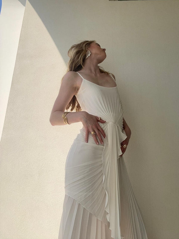 Model in a white dress wearing a floor length dress with delicate waterfall pleating, an open neckline with adjustable straps, and a slip on waist tie creating a flattering silhouette, standing against a light colored wall.