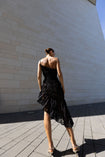 Model in a black sleeveless dress pleated in a sunray pattern with French baroque style lace standing against a beiModel