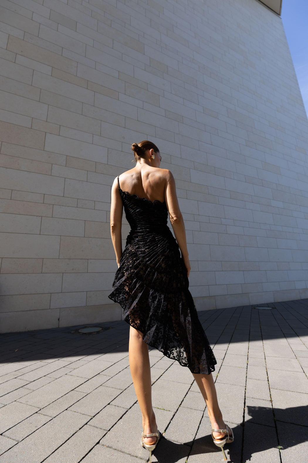 Model in a black sleeveless dress pleated in a sunray pattern with French baroque style lace standing against a beiModel