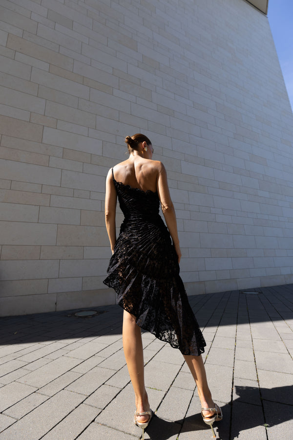 Model in a black sleeveless dress pleated in a sunray pattern with French baroque style lace standing against a beiModel