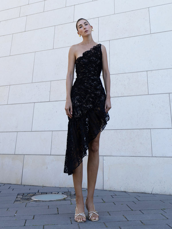 Model wearing a black coloured sleeveless dress pleated in a sunray pattern with French baroque style lace against a light-colored wall.