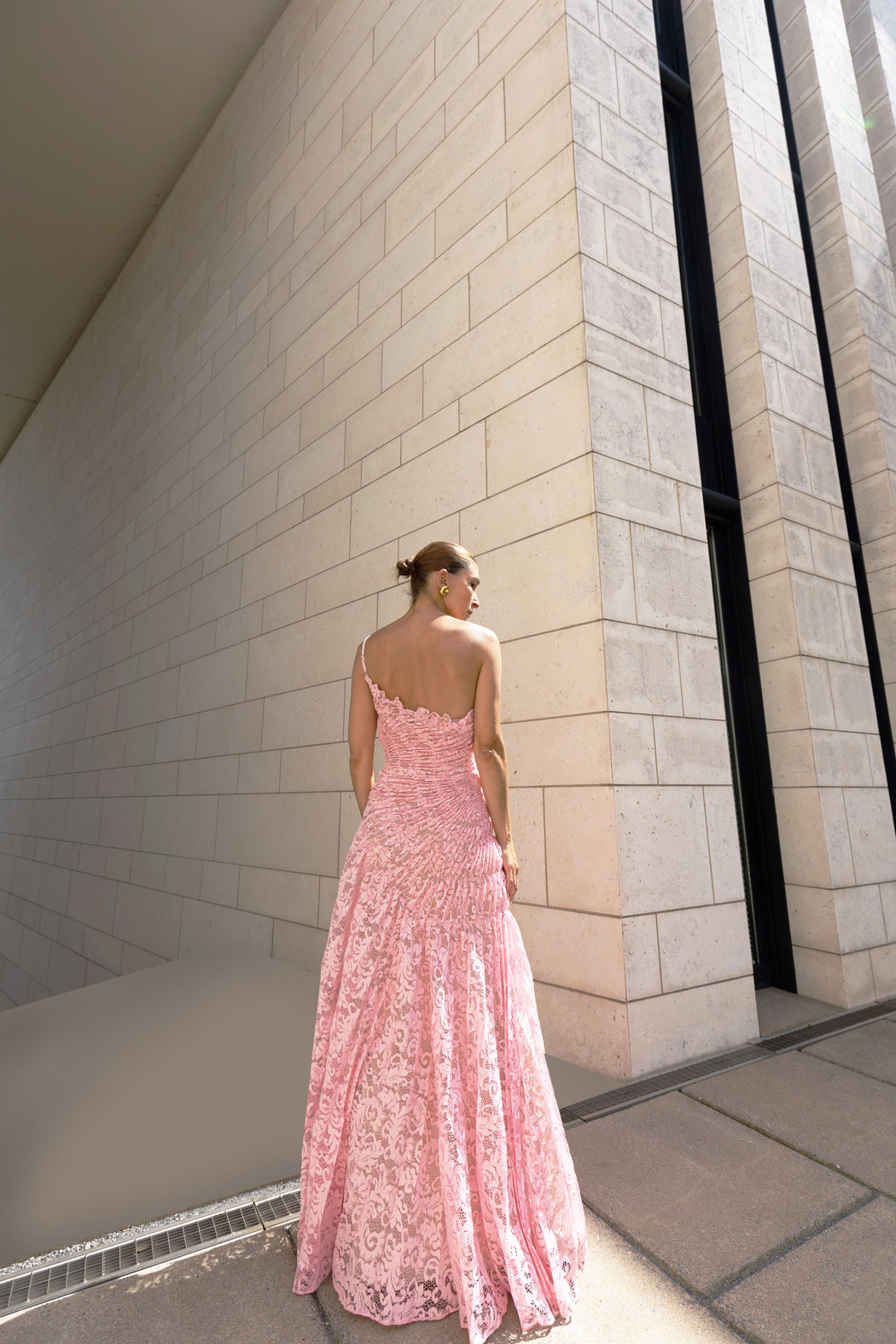 Model in pink sleeveless floor-length dress pleated in a sunray pattern with French baroque style lace facing a light-colored wall.