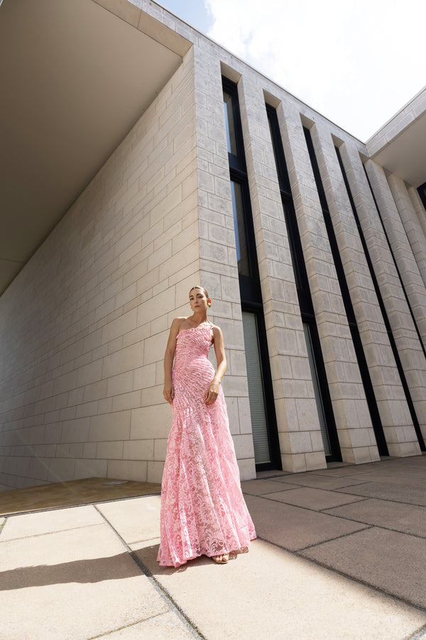 Model in pink sleeveless floor-length dress pleated in a sunray pattern with French baroque style lace standing outside a modern building.