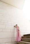 Model in pink sleeveless floor-length dress pleated in a sunray pattern with French baroque style lace standing on a staircase with a white tiled wall