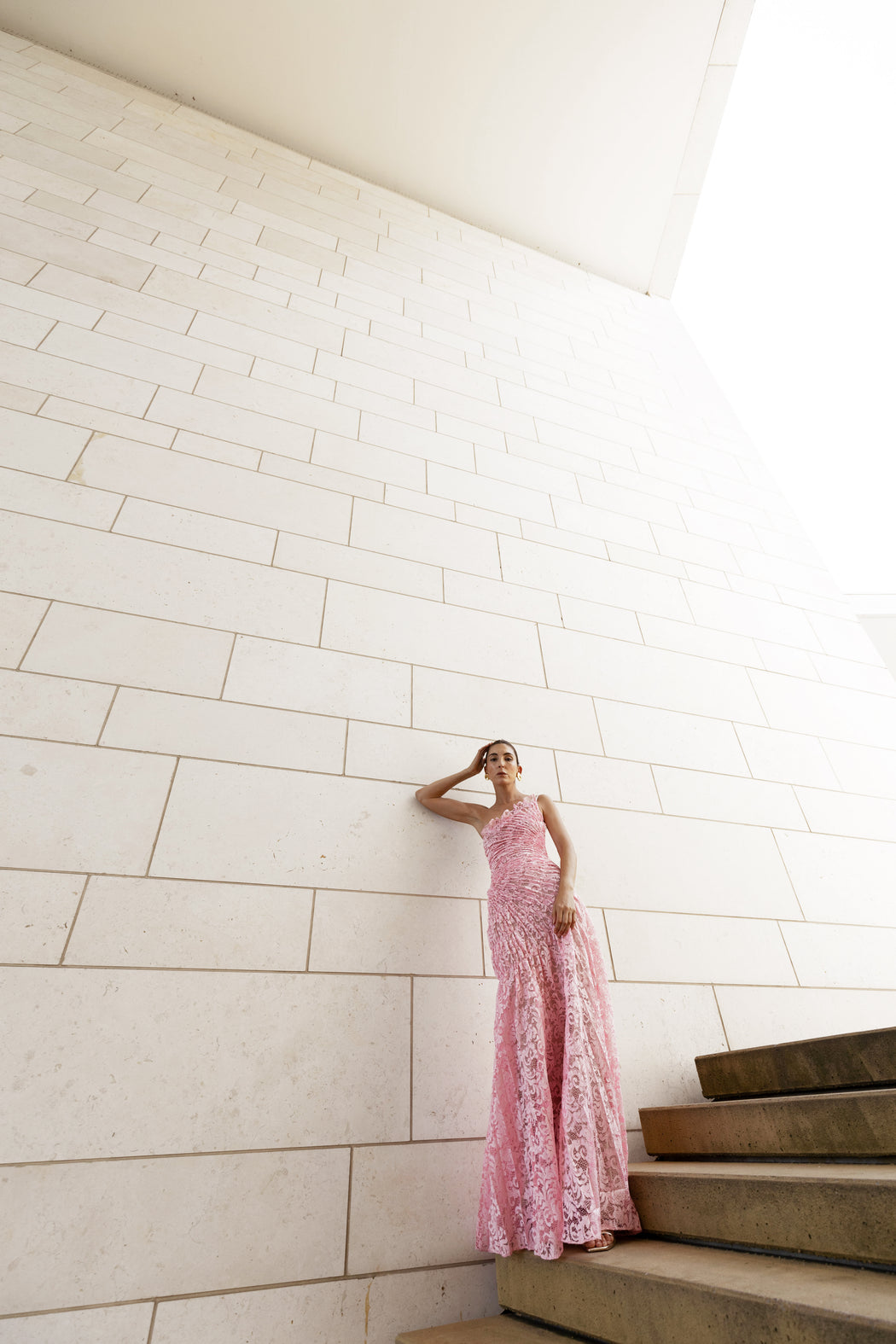 Model in pink sleeveless floor-length dress pleated in a sunray pattern with French baroque style lace standing on a staircase with a white tiled wall