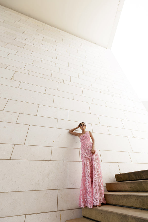 Model in pink sleeveless floor-length dress pleated in a sunray pattern with French baroque style lace standing on a staircase with a white tiled wall