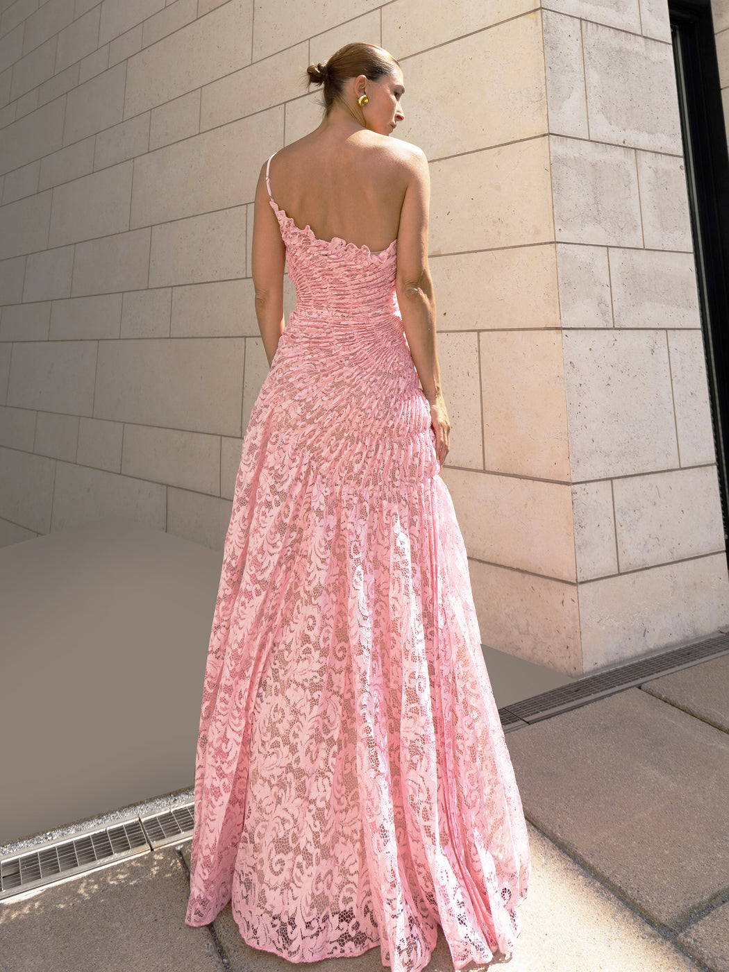 Model in pink sleeveless floor-length dress pleated in a sunray pattern with French baroque style lace standing against a light-coloured brick wall.