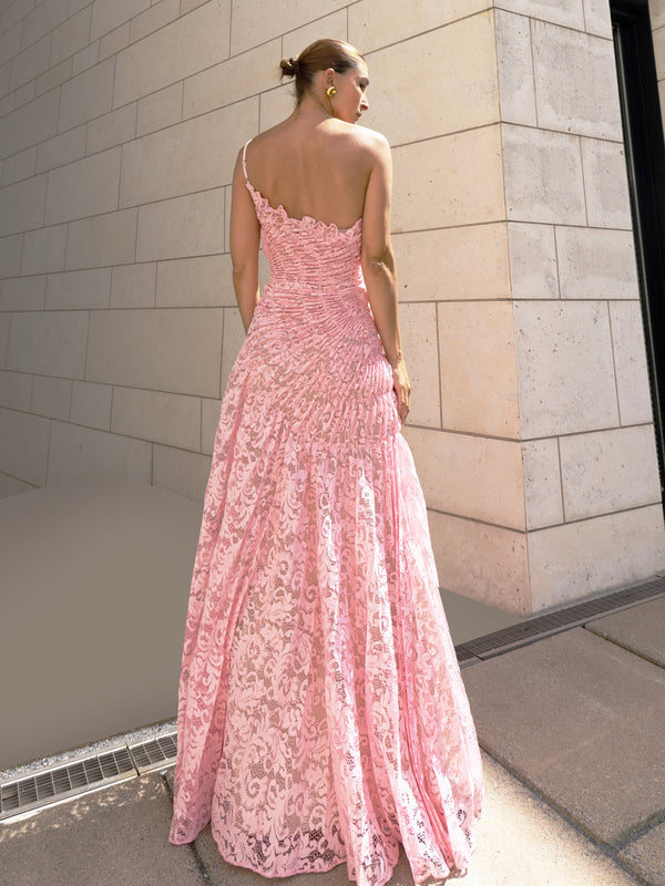 Model in pink sleeveless floor-length dress pleated in a sunray pattern with French baroque style lace standing against a light-coloured brick wall.