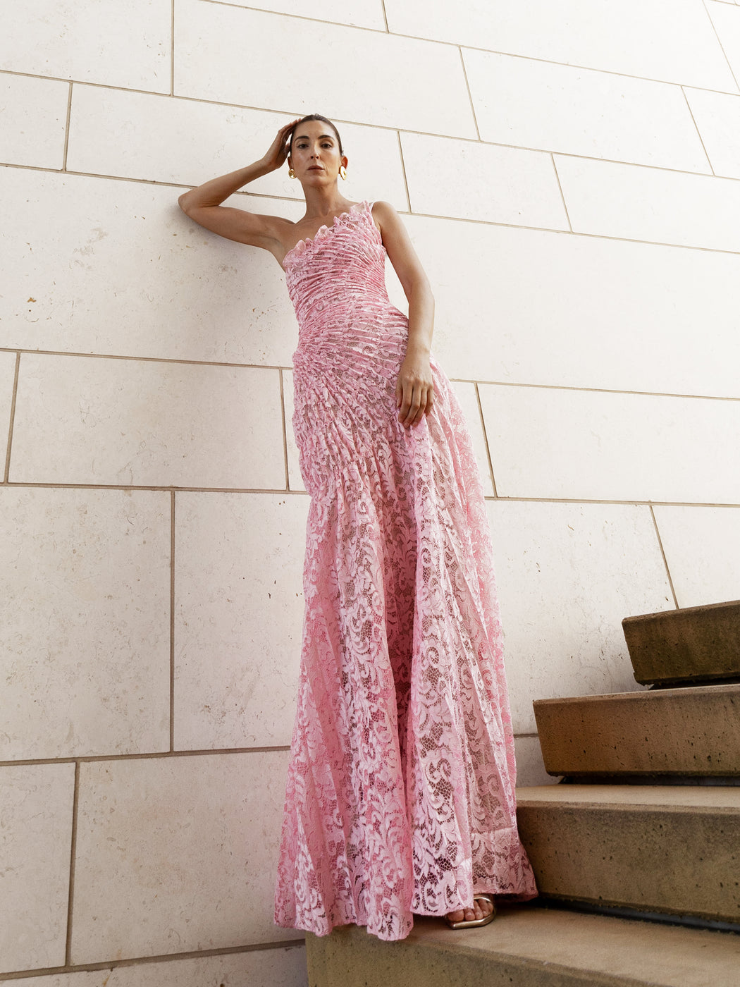 Model in pink sleeveless floor-length dress pleated in a sunray pattern with French baroque style lace standing on steps with a light-colored wall background